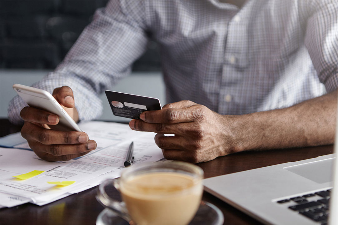 cropped shot of a mans hands holding a phone and payment card in front of a laptop 